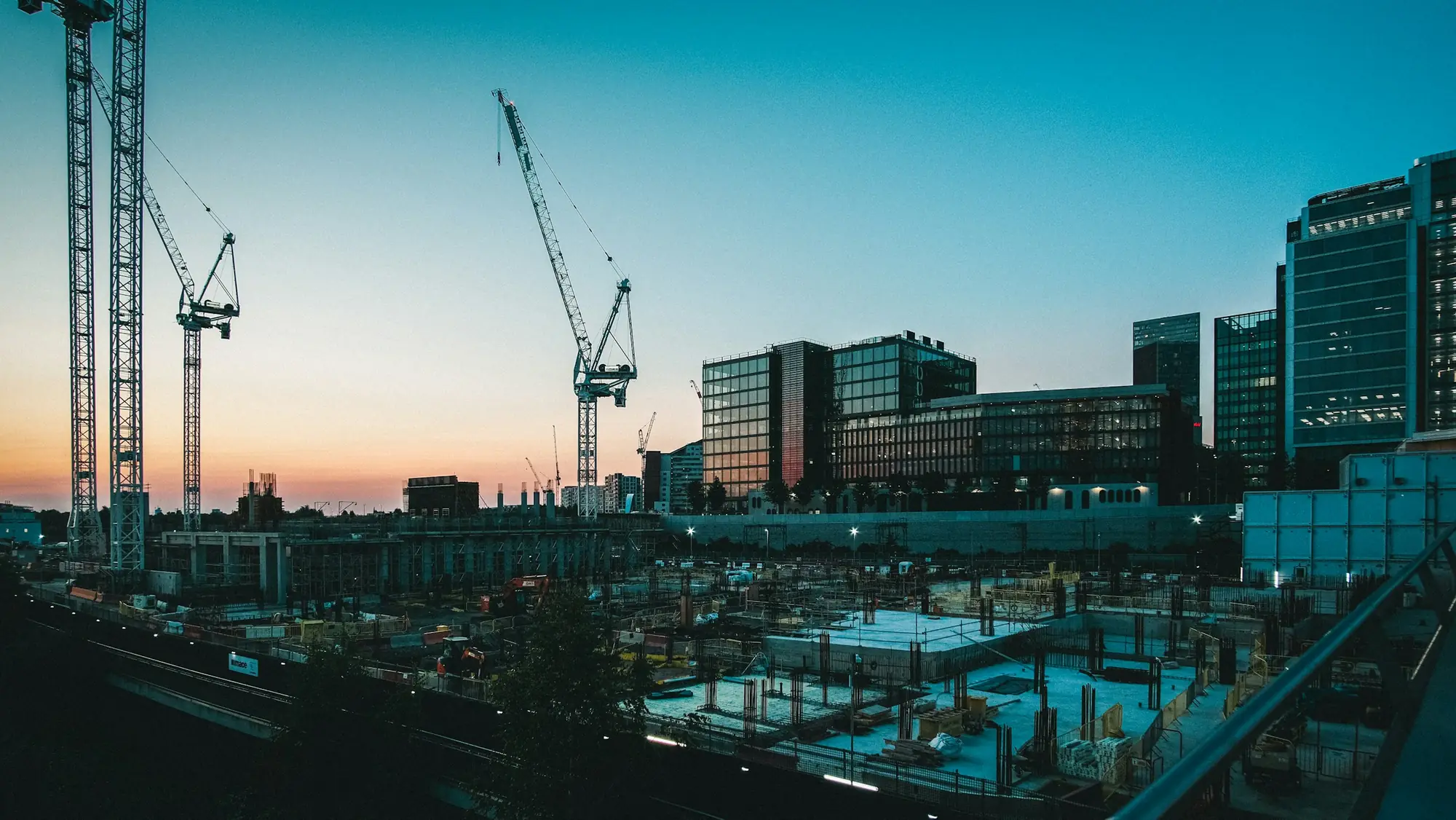 Tower cranes over a construction site at sunset, with modern office buildings behind